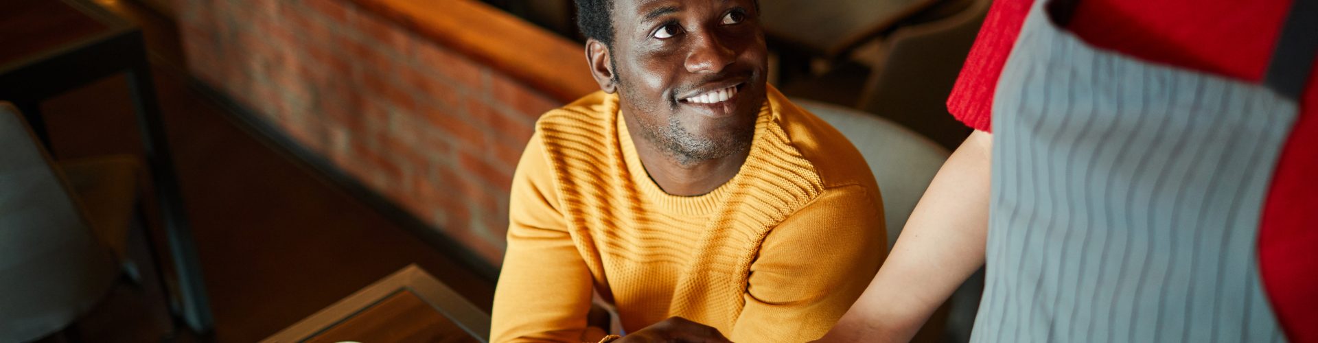 Young smiling man sitting by table and looking at waitress while paying for his order through smartphone