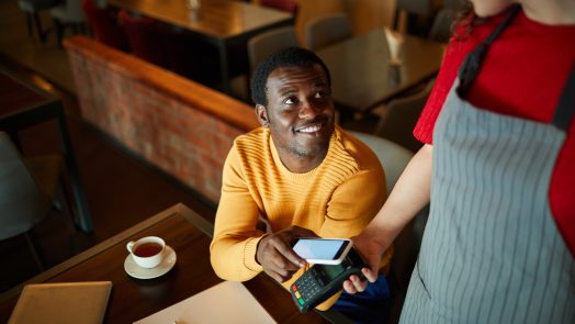 Young smiling man sitting by table and looking at waitress while paying for his order through smartphone