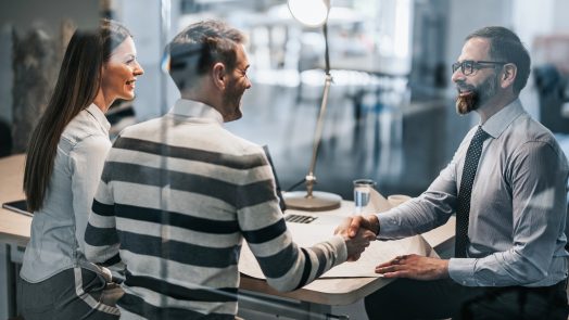 Happy real estate agent shaking hands with young couple after successful meeting in the office. The view is through glass.