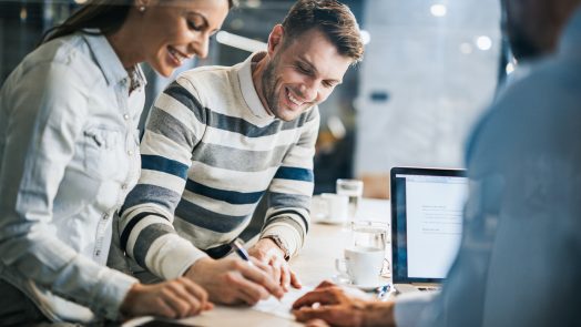 Young happy couple signing a document while being on a meeting with their real estate agent in the office. The view is through glass.
