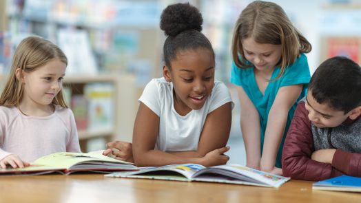 A group of friends are sitting at a table in their classroom. They are reading a book together as they smile and laugh.