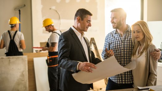 Young happy couple talking to their investor about housing plan inside of an apartment. Workers are in the background.