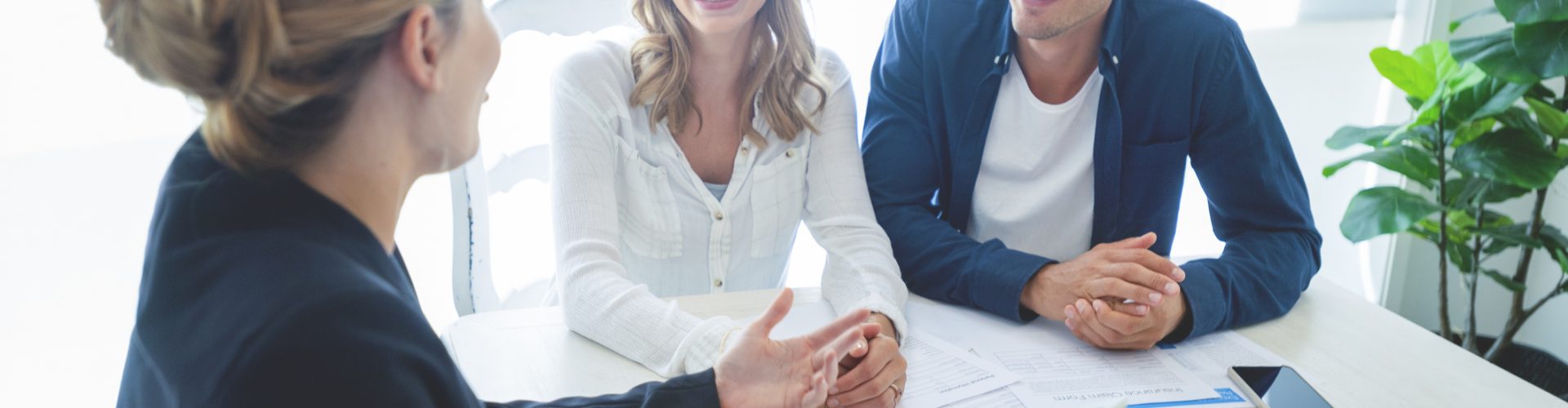 Insurance agent with couple looking through documents. The agent is holding a digital tablet. Couple are casually dressed. They sitting at a table at home and are looking a little happy and smiling.
