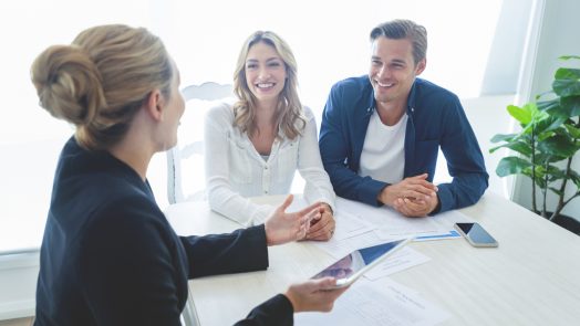 Insurance agent with couple looking through documents. The agent is holding a digital tablet. Couple are casually dressed. They sitting at a table at home and are looking a little happy and smiling.