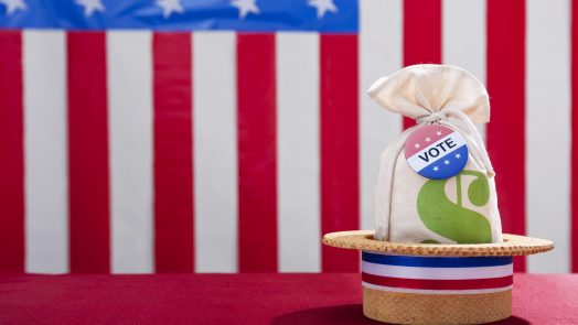 Political Rally Money and the Vote. A skimmer hat sitting on a table with a money bag and a Vote button against a large US flag in the background. Super PAC Money trying to buy vote that will influence the US Elections