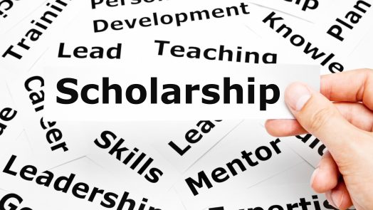 Hand holding a piece of paper with the word Scholarship printed on it above other related words in the background.