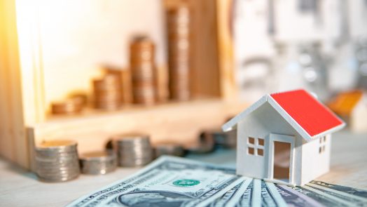 Property or real estate investment concept. Home mortgage loan rate. Saving money for future retirement. Miniature house model with stacked coins and dollar currency banknotes on wooden table.