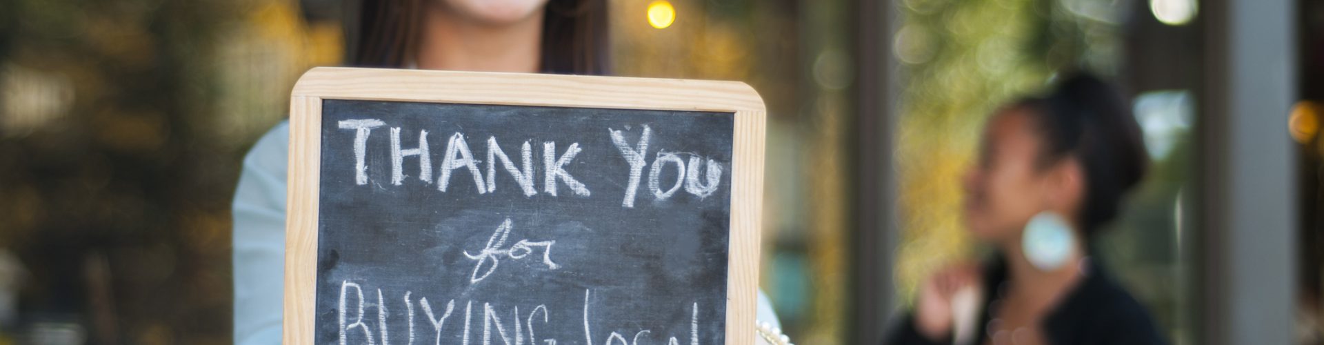 Young business owner holding a "Thank You for Buying Local" sign in front of her store with shopper in the background.