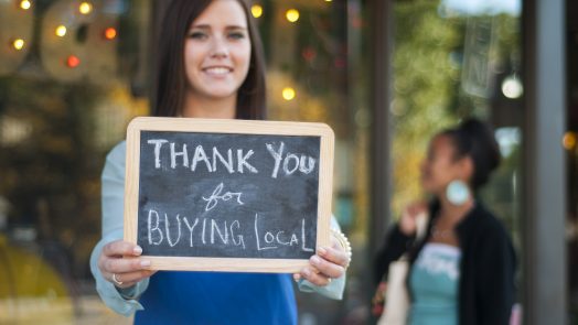 Young business owner holding a "Thank You for Buying Local" sign in front of her store with shopper in the background.