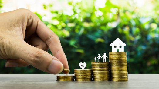 Saving or making money for family life in future concept. A man hand putting coin into rising stack of coins with heart, family and small house model on wood table. Depicts sustainable financial goal.