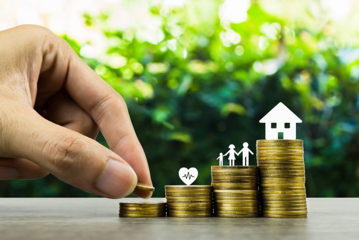 Saving or making money for family life in future concept. A man hand putting coin into rising stack of coins with heart, family and small house model on wood table. Depicts sustainable financial goal.